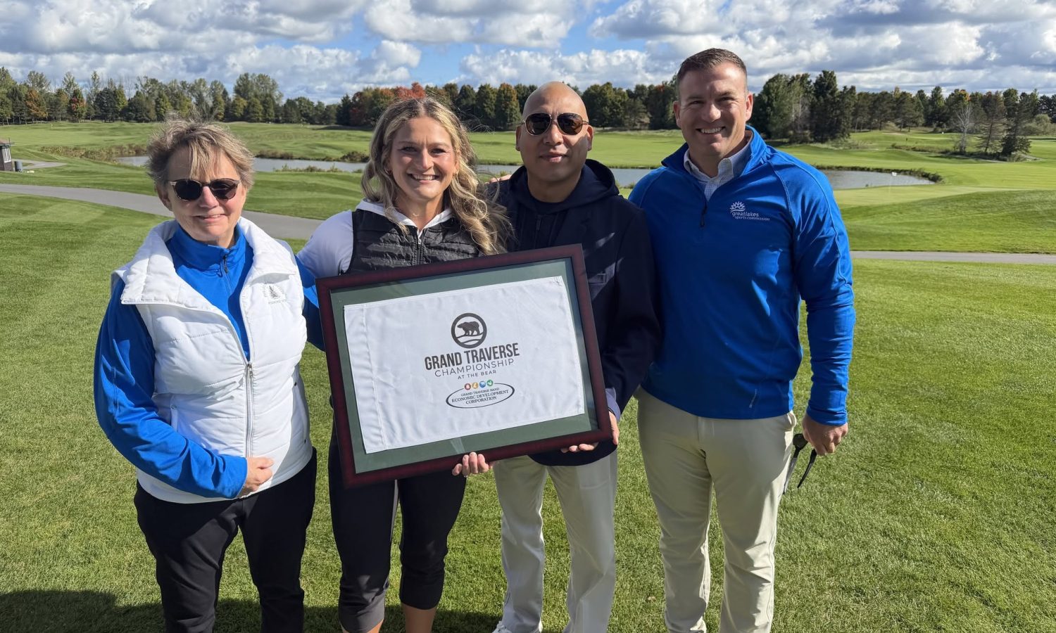 Four people on a golf course holding a framed Grand Traverse Championship at The Bear flag.